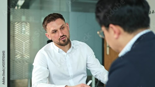 Two businessmen, one Caucasian with a beard and one Asian, sit and talk seriously during an important business meeting in a modern office.