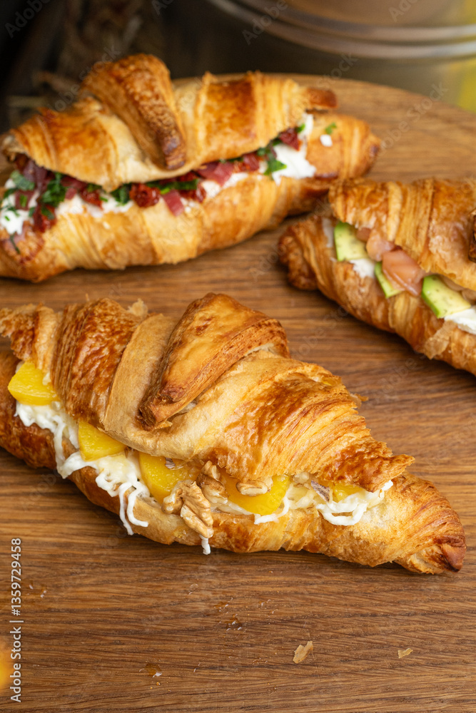 Three gourmet croissant sandwiches with different fillings on wooden board: peach and walnut, salmon and avocado, cured meat and herbs – rustic outdoor brunch Three gourmet croissant sandwiches with different fillings on wooden board: peach and walnut, salmon and avocado, cured meat and herbs – rustic outdoor brunch