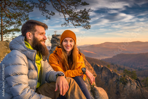 A bearded man and a woman in outdoor gear sit on a rocky peak at sunset. Both wear hiking watches, and the woman, in an orange beanie and jacket, looks at the camera with a warm smile.