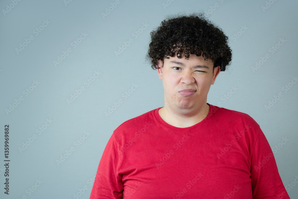 Young man frowning, looking upset. He wears red shirt and natural curly hair. Ideal for negative emotion, disappointment, frustration. Annoyed and bored face expression. Copy space on gray background. Young man frowning, looking upset. He wears red shirt and natural curly hair. Ideal for negative emotion, disappointment, frustration. Annoyed and bored face expression. Copy space on gray background.