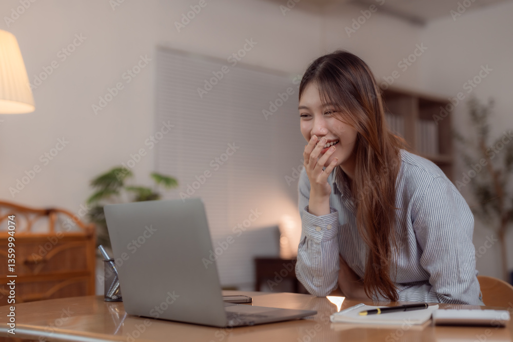 Young Asian businesswoman laughing while using a laptop in a cozy home office, enjoying online communication and watching funny videos during a break Young Asian businesswoman laughing while using a laptop in a cozy home office, enjoying online communication and watching funny videos during a break