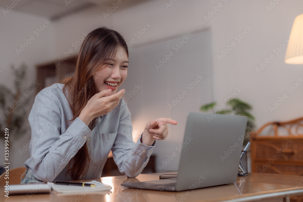 Young Asian businesswoman working from home, engaging in a lively video conference on her laptop, laughing and playfully pointing at the screen during the meeting Young Asian businesswoman working from home, engaging in a lively video conference on her laptop, laughing and playfully pointing at the screen during the meeting