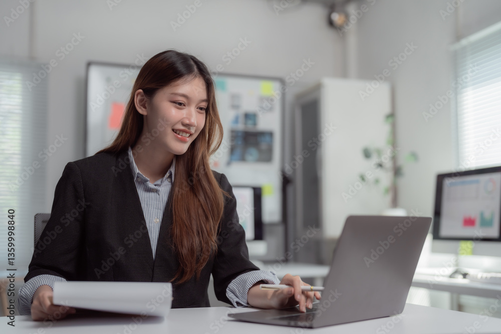 Young Asian businesswoman smiling while holding documents and working on a laptop at a modern office desk, analyzing financial reports with a professional demeanor Young Asian businesswoman smiling while holding documents and working on a laptop at a modern office desk, analyzing financial reports with a professional demeanor