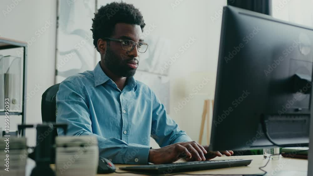 Serious African American man wearing glasses for better vision while sitting at his desk. Typing on keyboard while concentrating on computer screen. Focusing on important task or analyzing data. Serious African American man wearing glasses for better vision while sitting at his desk. Typing on keyboard while concentrating on computer screen. Focusing on important task or analyzing data.