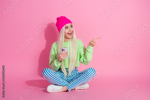 Young stylish woman in vibrant casual outfit sitting on pink background pointing cheerfully, holding smartphone