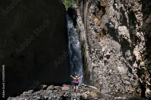 Obraz na płótnie A small silhouette of a woman in a red T-shirt standing with her arms raised in front of a waterfall in Arusha National Park, Tanzania w salonie