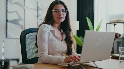 Charming Caucasian woman with brown hair and glasses sitting at desk and smiling while typing on laptop. Looking warm and approachable while working. Stylish office. Manager busy entering email.
