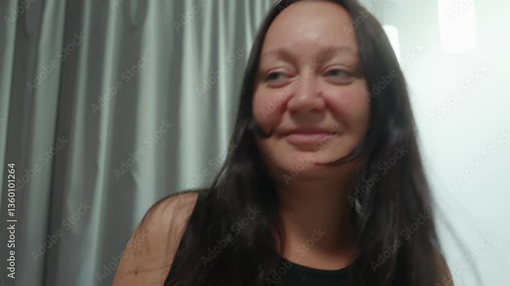 Energetic brunette woman dancing freely. Wearing sleeveless black shirt. Expressing pure joy and happiness while moving rhythmically in bright apartment interior with soft gray curtains