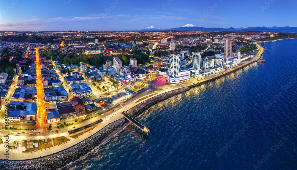 Naklejka premium Aerial drone view of Puerto Montt at twilight with a deep blue sky, faint orange sunset hues, and scattered clouds. The city glows with warm lights, and a serene waterfront, Osorno and Calbuco Volcano