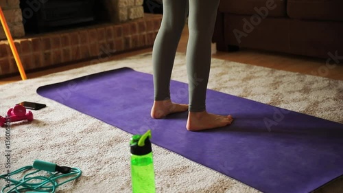 Close-up legs of young woman performing calf raises exercise on toes, yoga mat and fitness accessories in living room.