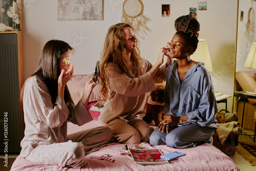 Wide shot of three biracial friends in pajamas sitting on bed and doing evening routine while having sleepover