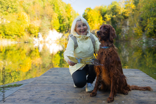 Happy senior 60s woman holds travel map searching location, with her best friend irish setter dog wear casual clothes in the autumn forest. Leisure, road trip, travel and people concept. 