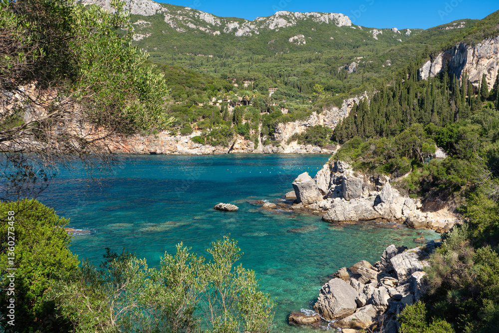 Sun shines to calm blue sea at Liapades bay, view from small rocky cliff near, green coniferous trees and bushes in foreground