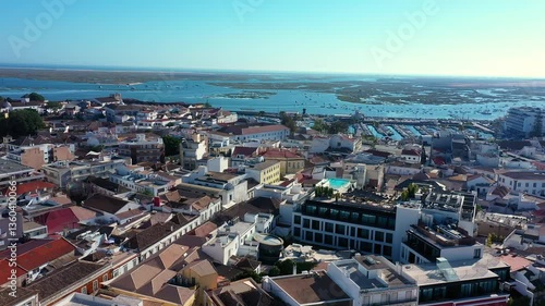 Aerial view of Faro, Portugal, displaying a mix of historic and modern architecture with red-tiled rooftops, a marina, and the Ria Formosa lagoon dotted with boats under a clear blue sky.