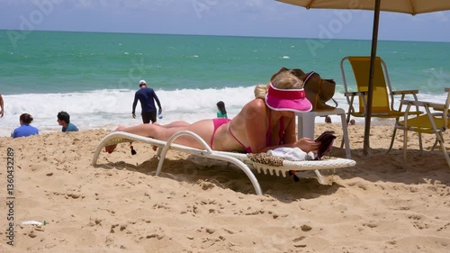 A woman in a pink bikini relaxes on a lounge chair at the beach, reading under the shade of an umbrella. The turquoise ocean waves crash in the background, creating a peaceful tropical scene.