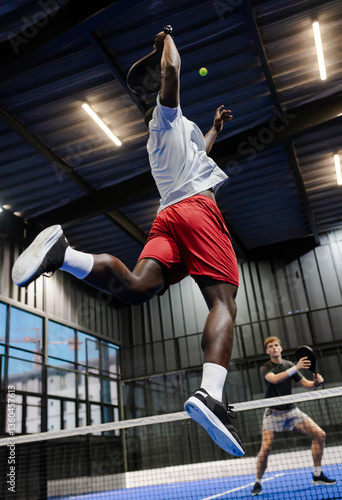 African American man jumping to hit ball during intense padel tennis match indoors