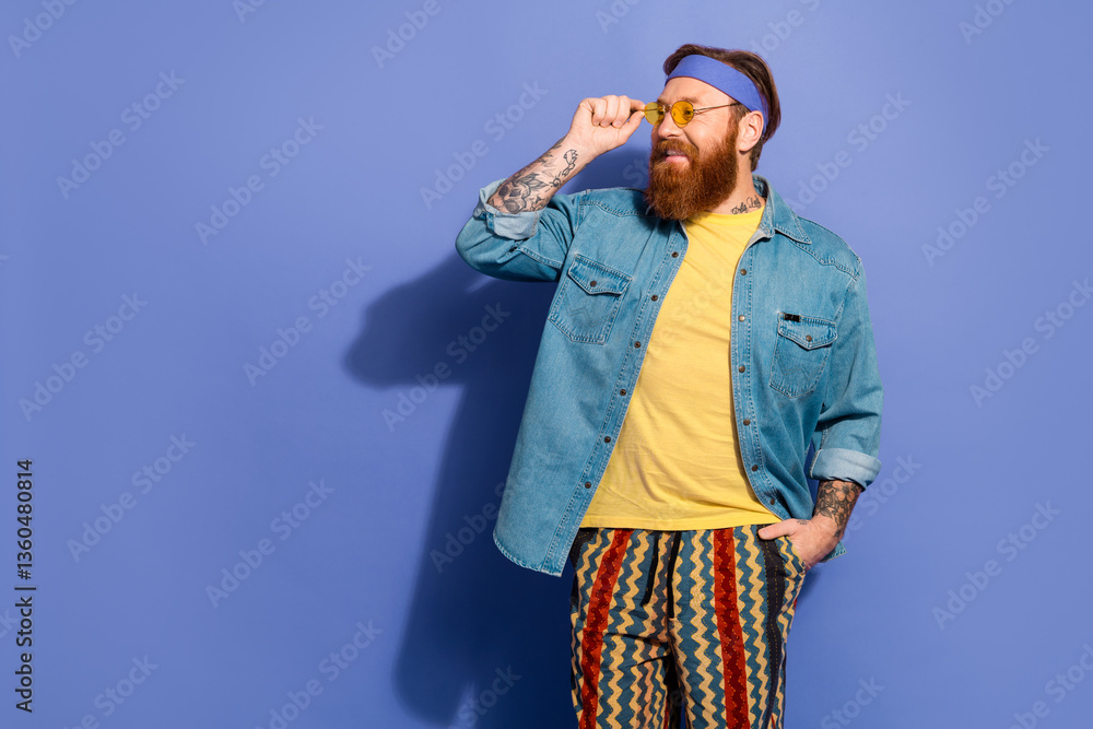 Fototapeta premium Trendy man with red beard wearing casual denim shirt and retro pants against a violet background, expressing happiness and confidence.