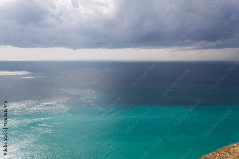 Fototapeta premium A stunning view of the seascape from a steep rocky cliff, the azure surface of the Black Sea and large cumulus clouds in the hot sultry summer in the Krasnodar region of the tourist city of Anapa.