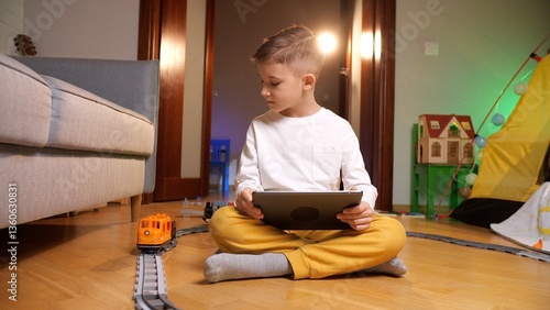 Happy boy in pajamas plays with a tablet and toy train in a cozy playroom, exploring technology and childhood games