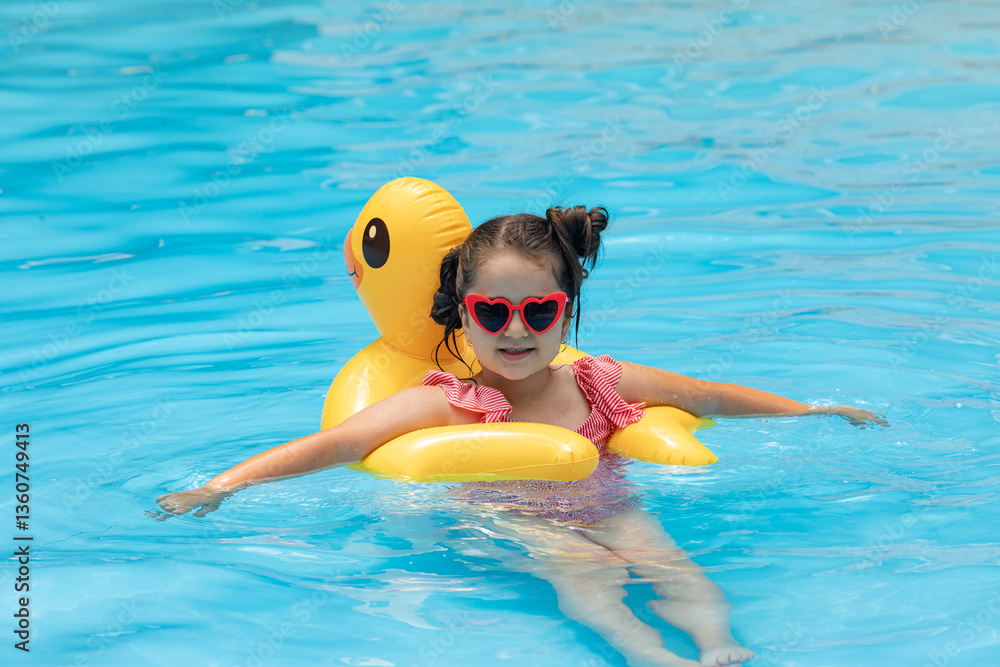 A little girl in sunglasses swims in an outdoor pool hugging an inflatable duckling. Sea holidays on vacation. Pretty female child smiles and swims along with inflatable yellow duck-shaped circle.