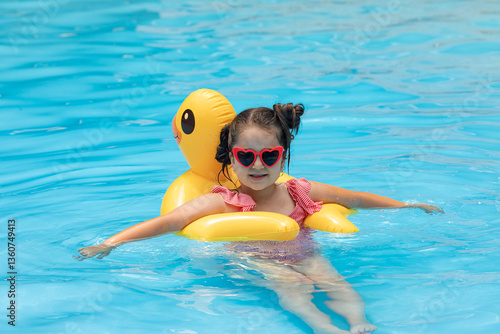 Obraz na płótnie A little girl in sunglasses swims in an outdoor pool hugging an inflatable duckling. Sea holidays on vacation. Pretty female child smiles and swims along with inflatable yellow duck-shaped circle. w salonie