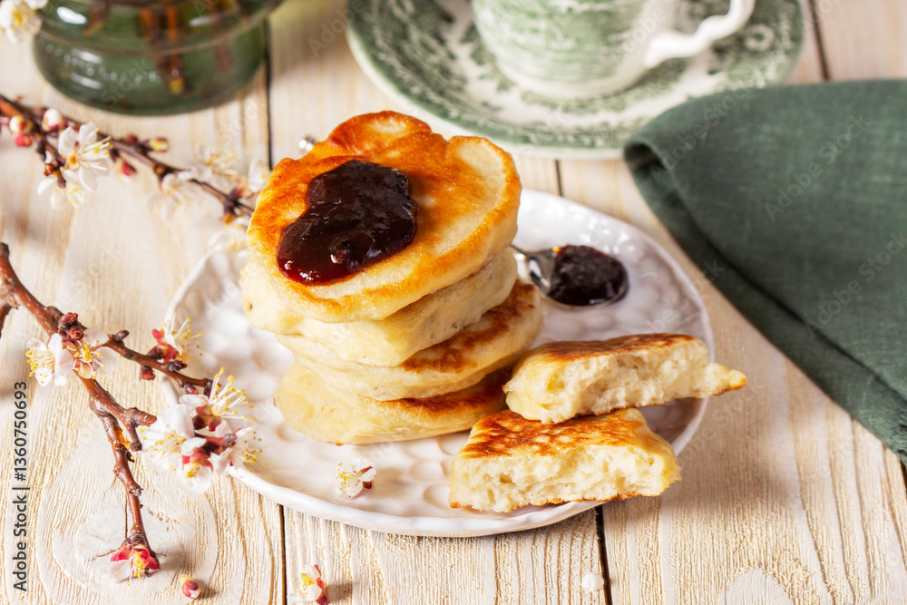 Lenten yeast pancakes with plum jam and tea, and a bouquet of flowering apricot branches on a light wooden table.