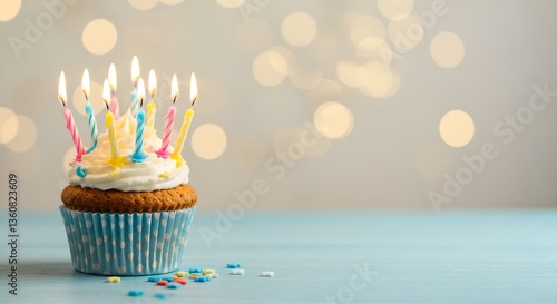 A delicious-looking cupcake with white frosting and seven lit candles in pink, yellow, and blue holders. The cupcake is in a  polka-dot paper cup, sitting on a light blue wooden surface.