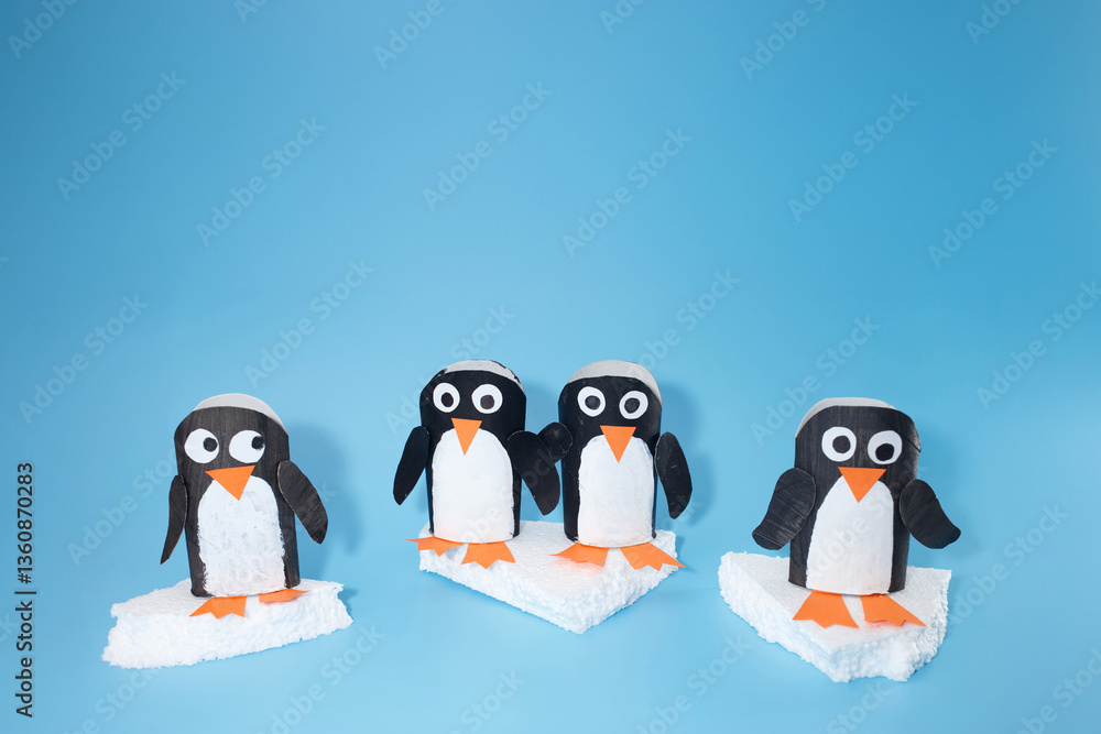 Obraz premium Penguin crafts using painted cardboard tubes with googly eyes, orange beaks, and feet on faux ice. Creative kids project on light blue surface for school activities, holiday decor, DIY inspiration