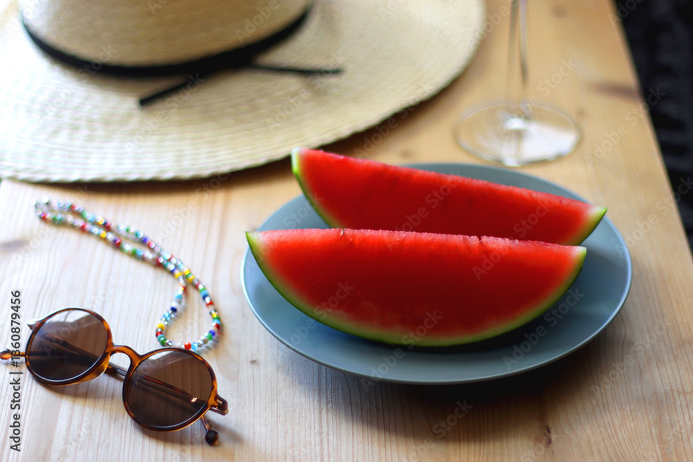 Plate with watermelon slices, glass of orange juice or cocktail, straw hat, colorful beaded necklace and sunglasses on the wooden table. Summer essentials concept. Selective focus.