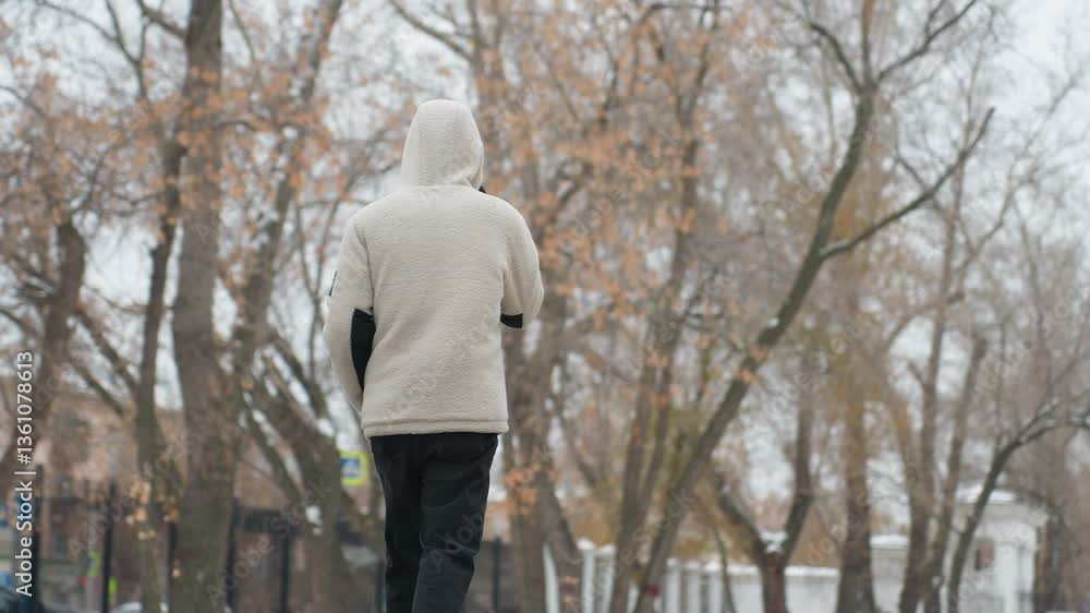 Back view of young adult in winter jacket walking and talking on phone with blurred background of dry trees, building, and unclear structures in urban environment on a cold day