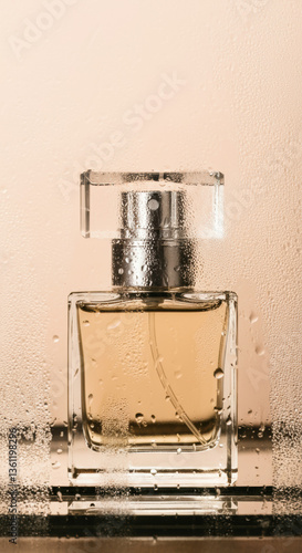 A sleek perfume bottle rests behind a glass pane covered in droplets, creating a sensual and refined aesthetic. The soft lighting and peach-toned background give the image a sophisticated and contempo