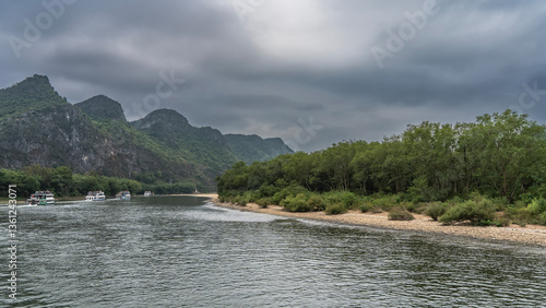Tourist ships sail along a beautiful river. Ripples, reflections on calm water in the riverbed. Green vegetation on the shore. Picturesque mountains against the sky and clouds. China. Li River.