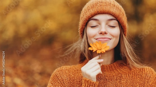 Young woman enjoying autumn outdoors with orange flower in hand wearing cozy sweater and knitted hat