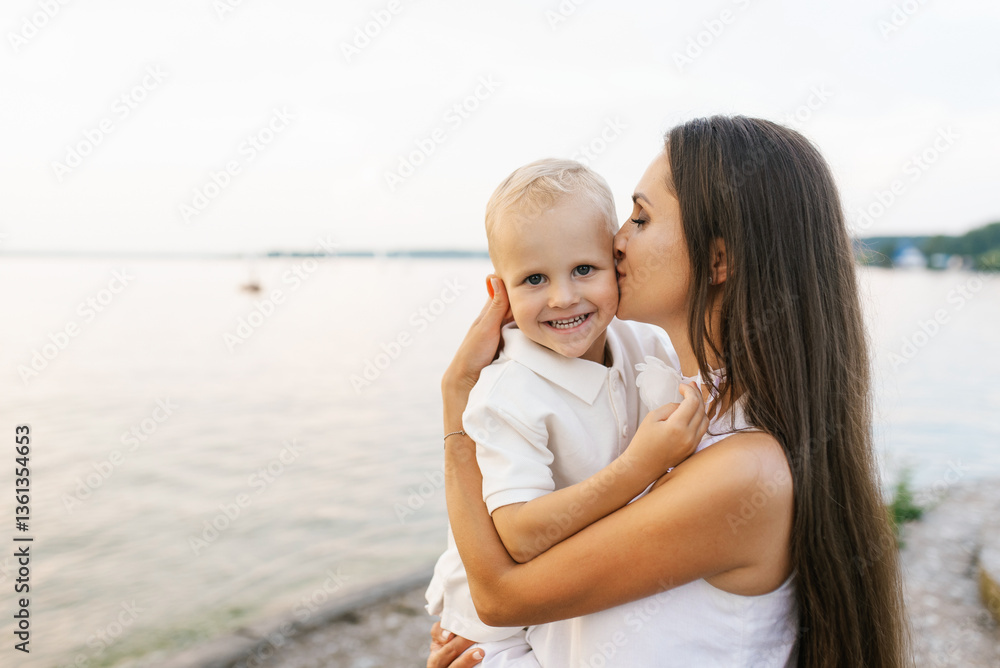 Mother is holding baby and kissing it on beach with water