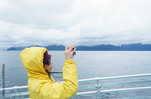 Woman in yellow raincoat taking smartphone photos on the cruise at Kenai Fjords National Park, Seward, Alaska.