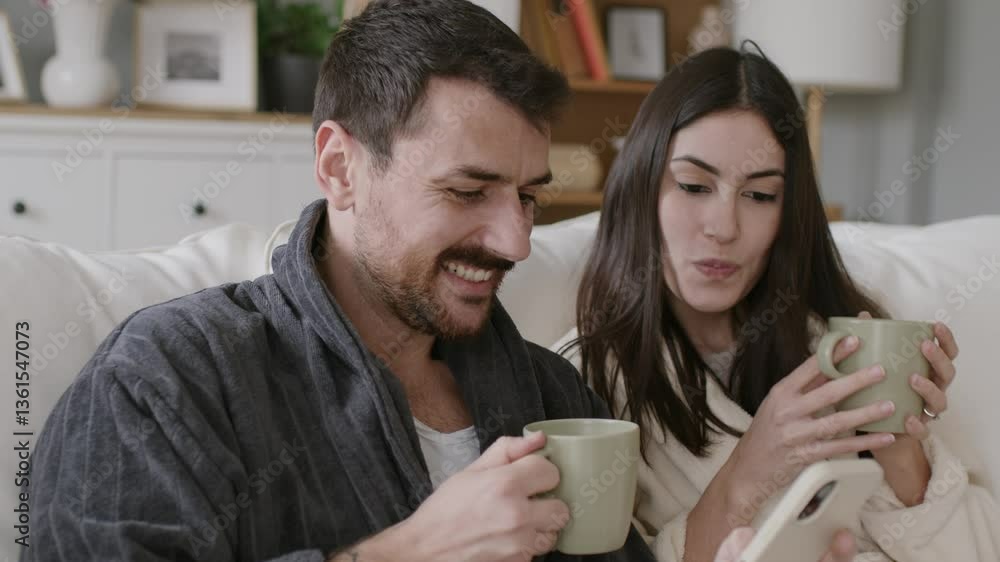 Couple wearing bathrobes laughing and drinking coffee while using smartphones on sofa. 4k video Couple wearing bathrobes laughing and drinking coffee while using smartphones on sofa. 4k video