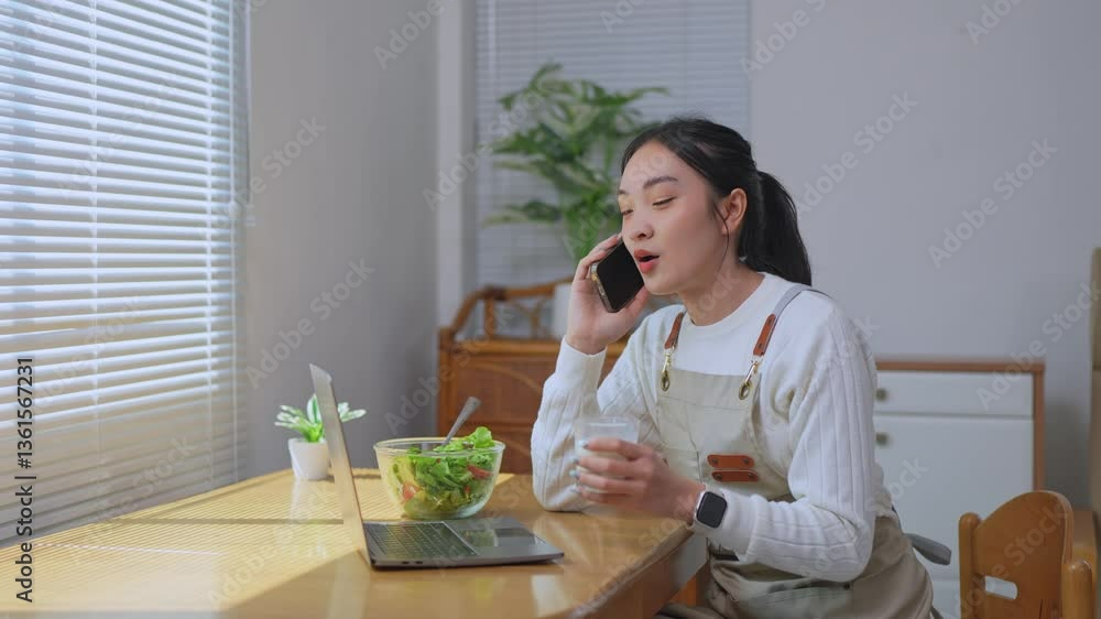 Young asian woman wearing apron enjoying lunch break at home, balancing work and relaxation while talking on phone and eating salad near laptop Young asian woman wearing apron enjoying lunch break at home, balancing work and relaxation while talking on phone and eating salad near laptop