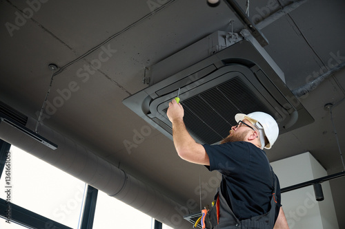Bearded man staying on ladder installing air conditioner at the office. Ceiling mounted cassette air conditioner in modern building. Loft style. Industrial interior design. Technician servicing