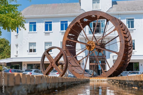 Rusty steel water wheel used to power grain mill on a display in center of Stellenbosch, south africa in the main city square. Sunny day, reflection in the water.