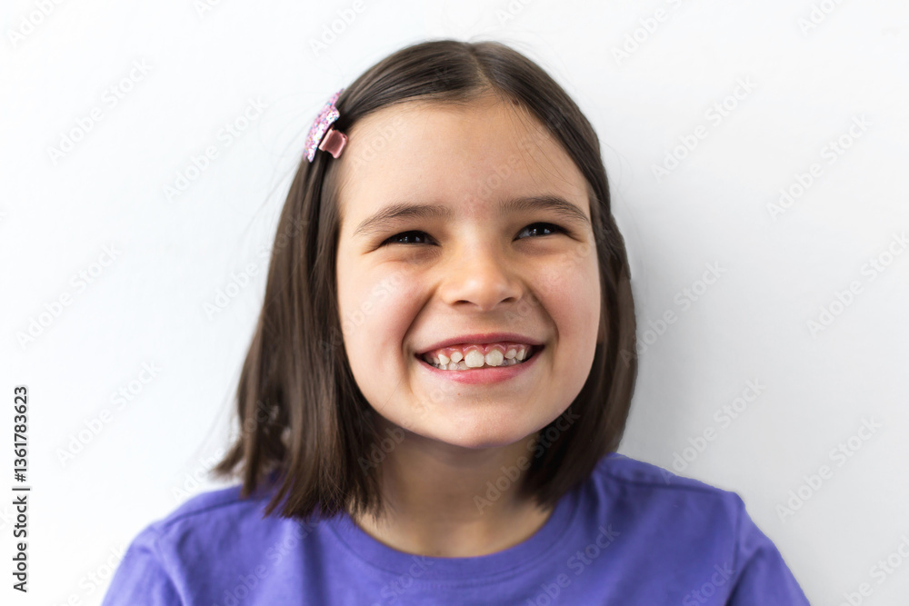 Close-up portrait of a happy 7-8-year-old girl with dark hair, wearing a purple shirt and a pink hair clip, smiling brightly and showing her teeth against a white background Close-up portrait of a happy 7-8-year-old girl with dark hair, wearing a purple shirt and a pink hair clip, smiling brightly and showing her teeth against a white background