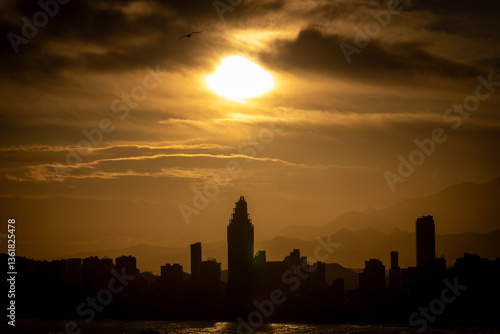 Photo of the sun setting in the evening over the old town of Benidorm in Spain showing the beautiful red sun set with clouds in the sky along side high rise apartments in the summer time