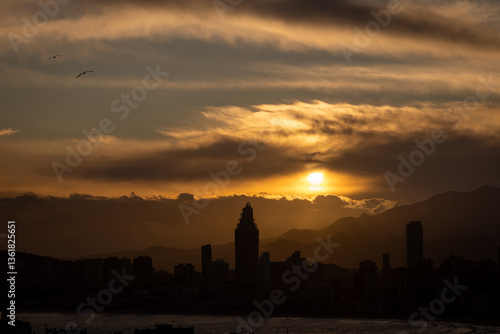 Photo of the sun setting in the evening over the old town of Benidorm in Spain showing the beautiful red sun set with clouds in the sky along side high rise apartments in the summer time