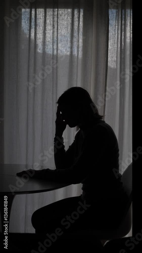 Vertical: Silhouette of a woman sitting at a table by the window, with her head in her hand in a gesture of despair, at night, with a gloomy atmosphere.