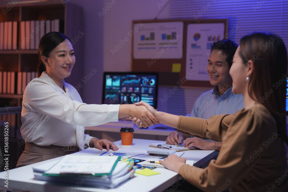 Obraz premium Two businesswomen shaking hands after reaching an agreement during a late-night meeting at the office, celebrating their successful collaboration with a colleague