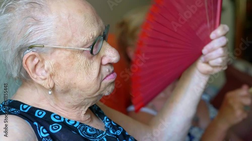A 90-year-old woman fans herself on a hot day, with another mature woman in the background doing the same. The scene highlights the intense heat and their efforts to stay cool.