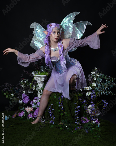 
full length portrait of pretty female model wearing fantasy elf costume with fairy wings and purple dress. sitting pose on magical dark studio background with romantic mossy flowers.