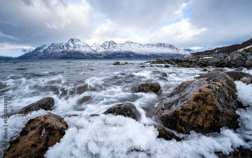 Waves crash against snow-covered rocks near a mountain range under a cloudy sky. The water is rough, and the scene is cold and dramatic Waves crash against snow-covered rocks near a mountain range under a cloudy sky. The water is rough, and the scene is cold and dramatic