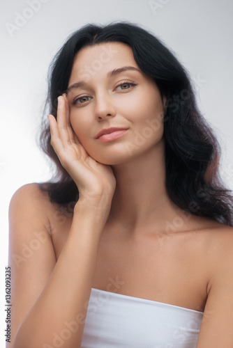 A young woman with radiant skin and dark hair gently touches her face, showcasing a natural skincare and beauty concept. Perfect for beauty, wellness, and cosmetic advertising purposes.