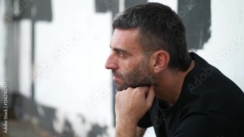 Vertical: A very sad and lonely young man with dark hair, sitting on a low wall in the park, side view, showing emotional distress and isolation.