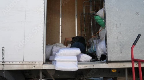 A truck driver with short hair organizes clean linens in the truck bed, preparing for the day's deliveries. He ensures the load is secure and ready for transport.